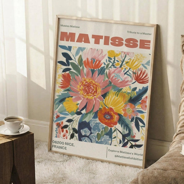 May include: A framed museum exhibition poster featuring vibrant watercolor-style flowers in coral, yellow, and blue tones against a cream background. The wooden frame leans against white paneled walls on a textured white rug, with a coffee cup on a wooden stool nearby and cozy fabric visible at the edge.