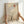 May include: Hands holding a light wood framed art print featuring a minimalist landscape with layered beige and tan mountains, a bare black tree silhouette, large pale circular moon, and small orange sun. The artwork has a desert-inspired color palette with soft neutral tones and geometric shapes creating depth through overlapping translucent layers.