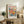 May include: Framed poster featuring vibrant floral artwork with coral and yellow blooms against cream background, displayed on rustic wooden sideboard next to fern plant, ceramic vase, and stacked books in neutral-toned living room setting.