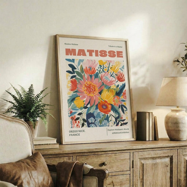 May include: Framed poster featuring vibrant floral artwork with coral and yellow blooms against cream background, displayed on rustic wooden sideboard next to fern plant, ceramic vase, and stacked books in neutral-toned living room setting.
