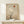 May include: Framed minimalist desert landscape artwork in natural wood frame, featuring bare black tree silhouette against layered beige and tan mountain ranges, large cream sun and small rust-colored circle in neutral sky, displayed on light wood surface with small white wrapped gift beside frame