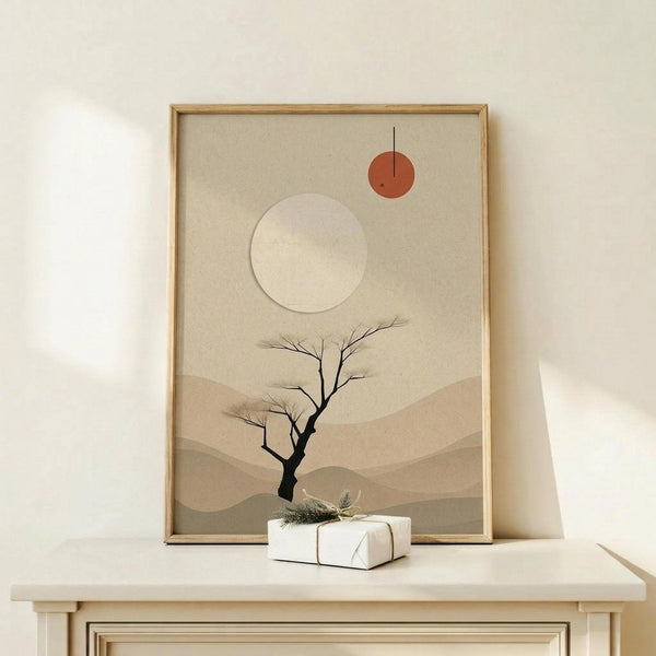 May include: Framed minimalist desert landscape artwork in natural wood frame, featuring bare black tree silhouette against layered beige and tan mountain ranges, large cream sun and small rust-colored circle in neutral sky, displayed on light wood surface with small white wrapped gift beside frame