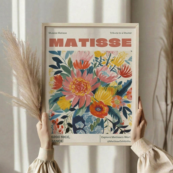 May include: Person holding a framed vintage-style floral poster featuring colorful painted flowers in coral, yellow, and blue tones. The museum-style print has "MATISSE" in bold red letters at the top and shows exhibition details at the bottom. Natural light filters through white curtains in the background, with dried wheat stems visible to the left. The person wears a cream-colored blouse with gathered sleeves.