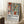 May include: Framed poster leaning against white wall corner featuring Matisse-inspired artwork with colorful vases, plants, and geometric shapes in warm oranges, greens, and blues. White text below reads "Celebrating Colors of Life" with descriptive paragraph. Small wooden table with white coffee cup visible on left, cream textured rug underneath, cozy room setting with natural window light.