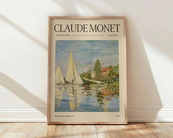 May include: Framed art print of Monet's impressionist painting showing sailboats with white sails on blue water, with green trees and buildings along the shoreline. The poster features "CLAUDE MONET EXHIBITION GALLERY" text at top and "Regattas at Argenteuil 1872" at bottom on cream background. Light wood frame sits against white wall on blonde hardwood floor with natural sunlight shadows.