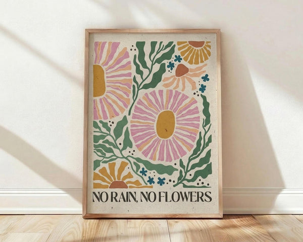 May include: Framed botanical poster print with colorful stylized flowers in pink, orange, and yellow with green foliage on cream background, featuring "NO RAIN, NO FLOWERS" text at bottom. Natural wood frame leaning against white wall on light wooden floor with soft sunlight shadows.