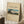 May include: Framed vintage-style poster print leaning against white wall corner on textured white rug. Features Japanese landscape artwork showing Mount Fuji, traditional thatched roof house, and lush greenery in muted earth tones. Cream background with black text and artist signature. Light wood frame complements cozy room setting with coffee cup on wooden block and beige knit throw blanket.