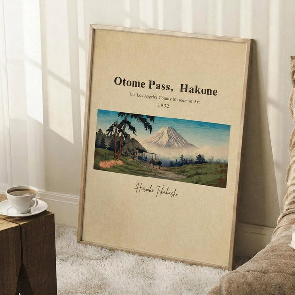 May include: Framed vintage-style poster print leaning against white wall corner on textured white rug. Features Japanese landscape artwork showing Mount Fuji, traditional thatched roof house, and lush greenery in muted earth tones. Cream background with black text and artist signature. Light wood frame complements cozy room setting with coffee cup on wooden block and beige knit throw blanket.