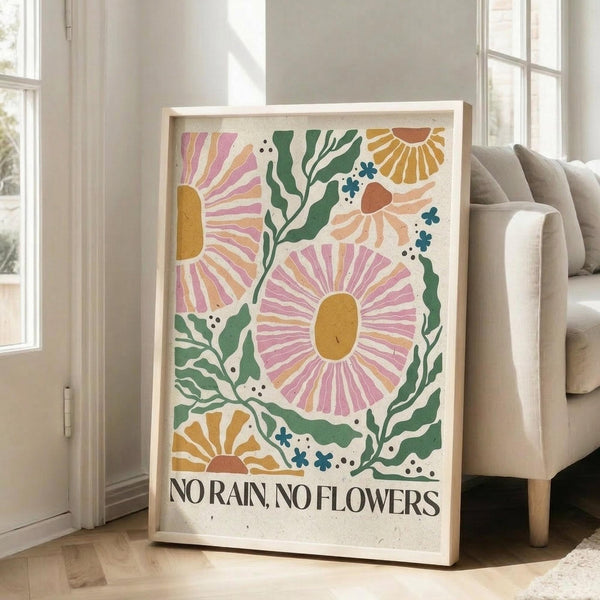 May include: Framed poster with colorful floral design featuring large pink and orange flowers with radiating petals, green leafy stems, and small blue accents on cream background. Black text reads "NO RAIN, NO FLOWERS" at bottom. Natural wood frame leans against white wall next to beige upholstered chair in bright living room with large windows.