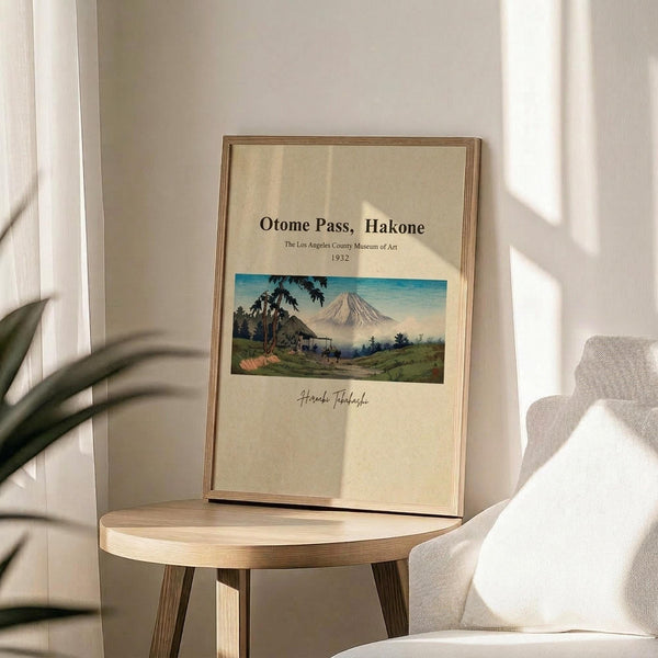 May include: Vintage Japanese landscape poster print in wooden frame, featuring Mount Fuji with traditional buildings and green hills in foreground, displayed on round wooden side table next to beige upholstered chair. Soft natural lighting creates gentle shadows across cream-colored wall. Green plant leaves visible in corner, creating serene minimalist home decor setting.