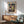 May include: Paul Klee abstract art print in wooden frame hanging above mid-century modern wooden credenza with white table lamp. Mint green upholstered armchair with wooden frame sits nearby. Poster features colorful geometric and organic shapes in blues, reds, yellows, and earth tones with intricate line work. Warm natural lighting creates shadows across white wall in minimalist living room setting.