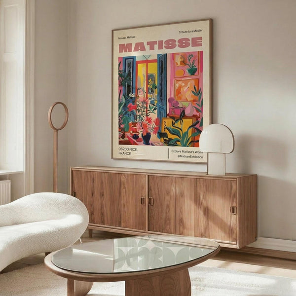 May include: Colorful Matisse exhibition poster in thin frame hanging above wooden credenza in modern living room. Poster features vibrant interior scene with plants, furniture, and windows in Matisse's distinctive style with red title text. Room has white curved chair, glass oval coffee table, floor lamp, and white table lamp on sideboard.