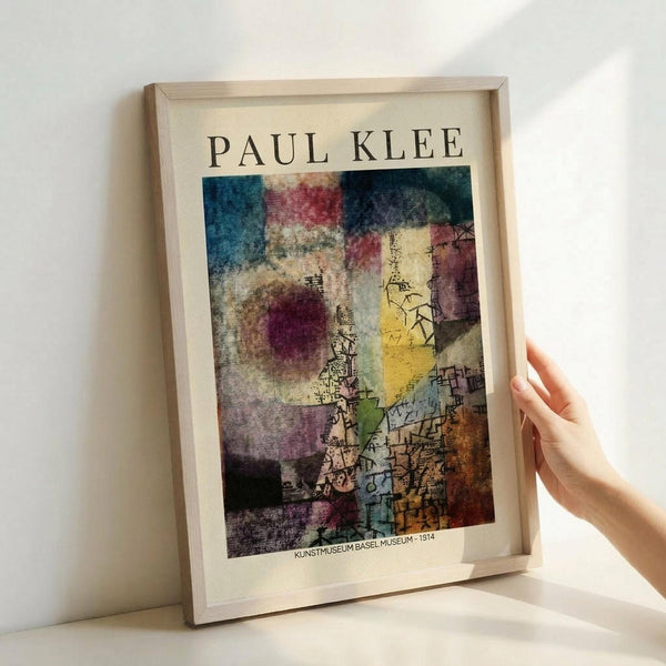 May include: Hands holding a framed art print featuring Paul Klee's abstract artwork with colorful geometric shapes, watercolor-like washes in blues, reds, yellows, and earth tones, plus intricate line drawings. The print shows "KUNSTMUSEUM BASEL - 1914" at bottom. Light wood frame against white background with soft natural lighting creating gentle shadows.