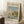 May include: Framed exhibition poster featuring Monet's impressionist sailing scene with white sailboats on reflective blue water, green trees and buildings along shoreline. Cream-colored poster with "CLAUDE MONET EXHIBITION" text at top, artwork title and 1872 date below painting. Light wood frame leaning against white wall corner, white coffee cup on wooden block nearby, beige textured rug and blanket visible.
