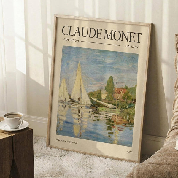 May include: Framed exhibition poster featuring Monet's impressionist sailing scene with white sailboats on reflective blue water, green trees and buildings along shoreline. Cream-colored poster with "CLAUDE MONET EXHIBITION" text at top, artwork title and 1872 date below painting. Light wood frame leaning against white wall corner, white coffee cup on wooden block nearby, beige textured rug and blanket visible.