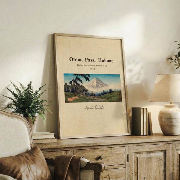 May include: Framed vintage Japanese landscape poster featuring Mount Fuji, rolling green hills, and traditional buildings in soft blues and greens. The cream-colored print sits on a rustic wooden sideboard next to books, a ceramic vase, and a potted fern in a neutral-toned living room setting.