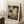 May include: Framed Paul Klee abstract art print leaning against white wall in cozy room setting. The artwork features vibrant watercolor-style composition with geometric and organic forms in blues, purples, yellows, and earth tones. Simple light wood frame surrounds cream-colored mat. Scene includes white coffee cup on wooden block and soft beige textiles on white fluffy rug, creating warm minimalist home decor atmosphere.
