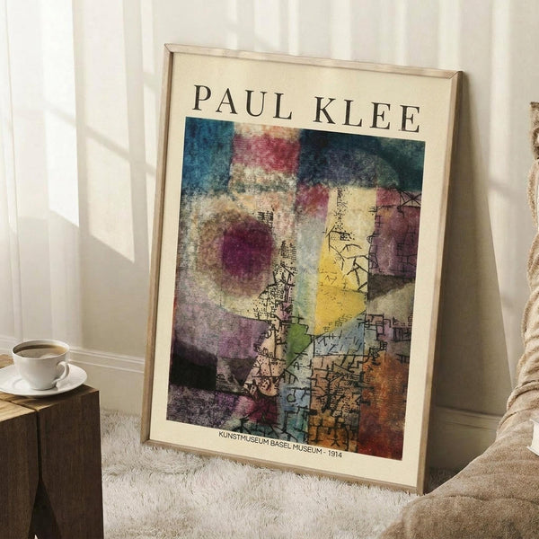 May include: Framed Paul Klee abstract art print leaning against white wall in cozy room setting. The artwork features vibrant watercolor-style composition with geometric and organic forms in blues, purples, yellows, and earth tones. Simple light wood frame surrounds cream-colored mat. Scene includes white coffee cup on wooden block and soft beige textiles on white fluffy rug, creating warm minimalist home decor atmosphere.
