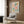 May include: Framed botanical poster with colorful stylized flowers in pink, orange, and green with "No Rain, No Flowers" text, displayed above a beige upholstered armchair with geometric patterned pillow. Modern living room setting features gold floor lamp, dark round side table with open book, wood panel wall, and white curtains.