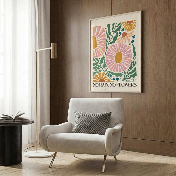May include: Framed botanical poster with colorful stylized flowers in pink, orange, and green with "No Rain, No Flowers" text, displayed above a beige upholstered armchair with geometric patterned pillow. Modern living room setting features gold floor lamp, dark round side table with open book, wood panel wall, and white curtains.