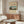 May include: Vintage Japanese landscape poster featuring Mount Fuji, traditional buildings, and green countryside in muted colors, displayed in thin brown frame above mid-century walnut credenza. Room shows cream walls, white modern chair, glass oval coffee table, and warm natural lighting creating soft shadows on furniture.