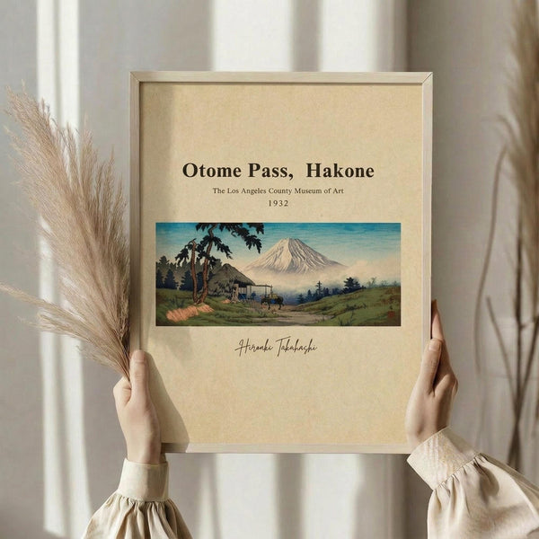 May include: Hands holding a framed vintage Japanese landscape print showing Mount Fuji, traditional buildings, and lush greenery in muted colors on cream background. Natural wooden frame, soft lighting, pampas grass visible to left, neutral beige clothing and curtains in background.