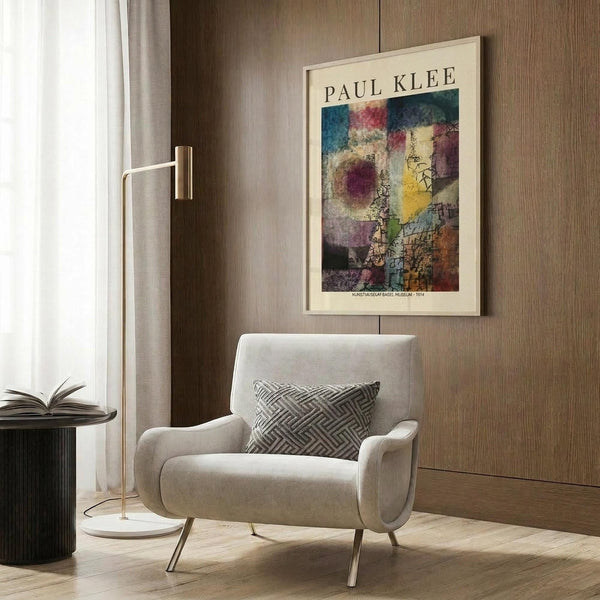 May include: Paul Klee abstract art poster with colorful geometric squares and circular forms in blues, reds, yellows and earth tones, displayed in light wood frame on textured wood wall above modern beige armchair with geometric patterned pillow, next to brass floor lamp and dark round side table with open book