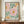 May include: Framed botanical art print with colorful abstract flowers in pink, orange, and yellow with green foliage on cream background, displaying "NO RAIN, NO FLOWERS" text at bottom. Print sits on wooden sideboard next to white ceramic vase and books in elegant room with beige walls and white trim molding.