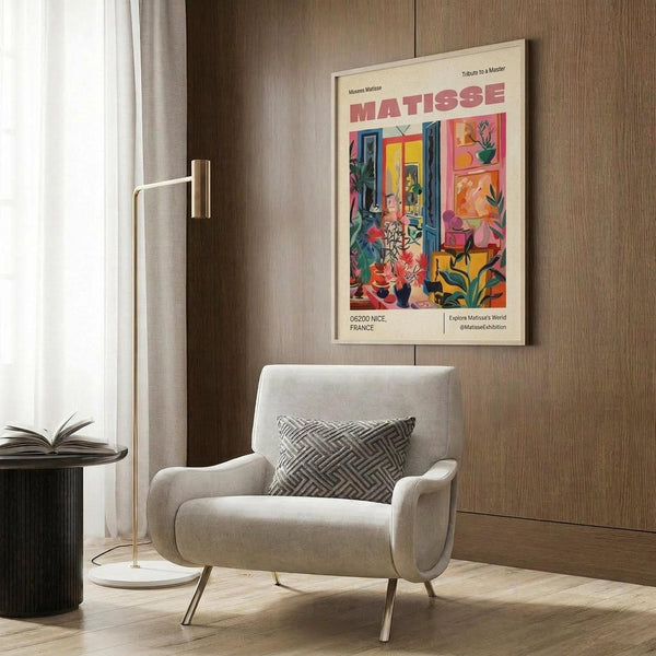 May include: Colorful Matisse exhibition poster in light wood frame hanging on wood-paneled wall above cream upholstered armchair with geometric pillow. Poster features vibrant interior scene with plants, furniture, and bold colors in pink, blue, yellow, and green. Modern reading nook with brass floor lamp and side table.