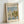 May include: Framed exhibition-style poster featuring Monet's sailboat painting with white sails on blue water, green trees and buildings in background. Light wood frame, cream matting with "CLAUDE MONET EXHIBITION GALLERY" text at top. Poster shows "Regattas at Argenteuil 1872" at bottom. Frame leans against white wall corner, casting subtle shadow.