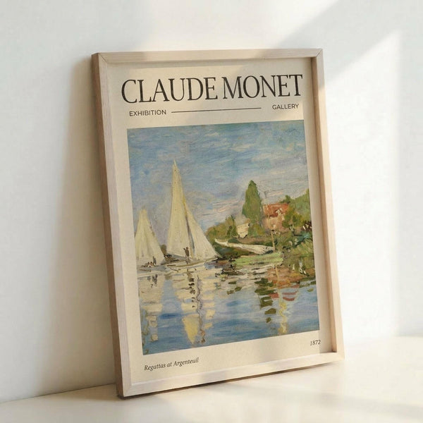 May include: Framed exhibition-style poster featuring Monet's sailboat painting with white sails on blue water, green trees and buildings in background. Light wood frame, cream matting with "CLAUDE MONET EXHIBITION GALLERY" text at top. Poster shows "Regattas at Argenteuil 1872" at bottom. Frame leans against white wall corner, casting subtle shadow.