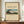 May include: Framed vintage Japanese landscape print showing Mount Fuji with rural village scene, displayed on wooden credenza with white ceramic vase and books. Cream matting surrounds colorful artwork featuring snow-capped mountain, traditional buildings, and green countryside under blue sky. Brown wooden frame against white paneled wall.