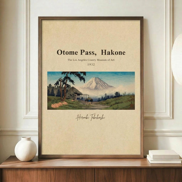 May include: Framed vintage Japanese landscape print showing Mount Fuji with rural village scene, displayed on wooden credenza with white ceramic vase and books. Cream matting surrounds colorful artwork featuring snow-capped mountain, traditional buildings, and green countryside under blue sky. Brown wooden frame against white paneled wall.