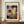 May include: Framed Paul Klee abstract art poster with vibrant watercolor-style patches in blues, reds, yellows, and earth tones, featuring geometric line drawings and text reading "KUNSTMUSEUM BASEL MUSEUM - 1914". The brown wooden frame sits on a light wood credenza beside a small white ceramic vase and stack of books against cream paneled walls.