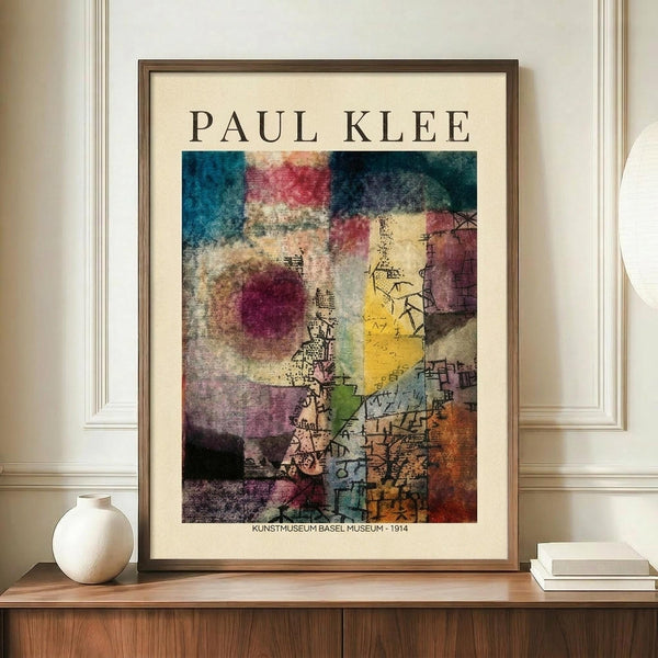 May include: Framed Paul Klee abstract art poster with vibrant watercolor-style patches in blues, reds, yellows, and earth tones, featuring geometric line drawings and text reading "KUNSTMUSEUM BASEL MUSEUM - 1914". The brown wooden frame sits on a light wood credenza beside a small white ceramic vase and stack of books against cream paneled walls.