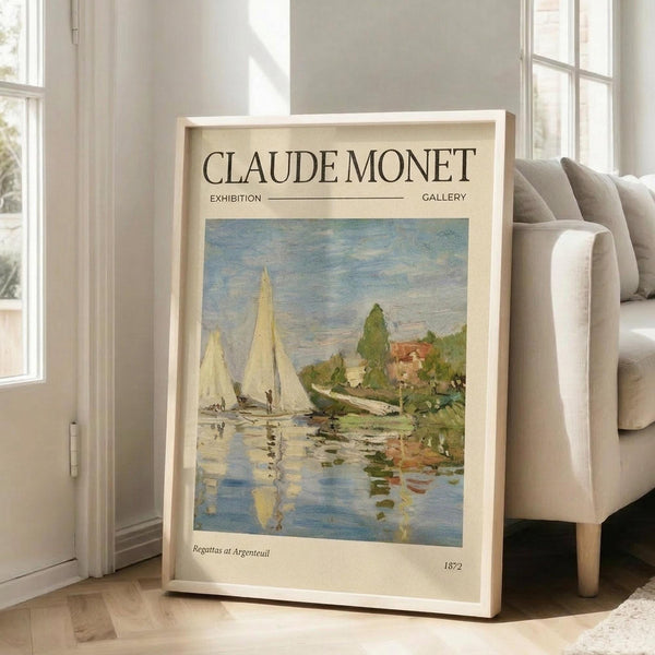 May include: Framed exhibition poster featuring Claude Monet's impressionist painting of white sailboats on reflective water with green trees and buildings along the shoreline. The cream-colored poster displays "CLAUDE MONET EXHIBITION GALLERY" at top with "Regattas at Argenteuil 1872" at bottom. Light wood frame leans against white wall next to beige upholstered chair on light hardwood flooring in bright, sunlit room.