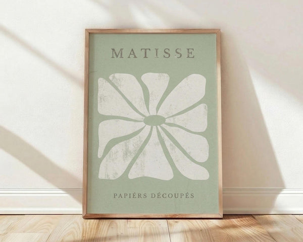 May include: Framed art print with sage green background featuring a white stylized flower with eight rounded petals radiating from center circle. Text reads "MATISSE" at top and "PAPIERS DÉCOUPÉS" at bottom in gray letters. Natural wood frame sits on light wooden floor against white wall with soft natural lighting creating diagonal shadows.