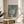 May include: Framed art print with stylized tree design featuring dotted green and blue foliage on blue background with yellow circular elements, displayed on light wood round side table next to beige upholstered chair with knit throw pillow, plant leaves visible in corner, natural window lighting creating soft shadows on neutral wall