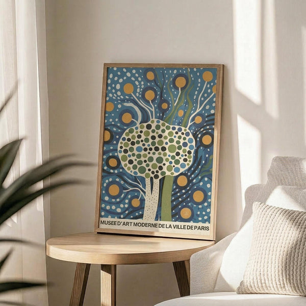 May include: Framed art print with stylized tree design featuring dotted green and blue foliage on blue background with yellow circular elements, displayed on light wood round side table next to beige upholstered chair with knit throw pillow, plant leaves visible in corner, natural window lighting creating soft shadows on neutral wall
