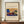 May include: A framed art print displayed on a wooden sideboard featuring Kandinsky's abstract artwork with vibrant blues, yellows, and reds in an expressionist landscape style. The poster has cream matting with "Painting with Troika" and "The Art Institute of Chicago" text above the artwork, and "Wassily Kandinsky" below. The brown wooden frame sits beside a small white ceramic vase and stack of books against white wainscoting walls.