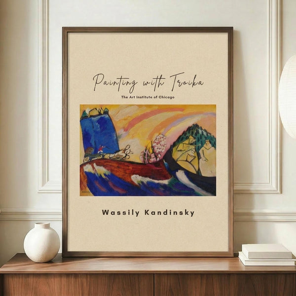 May include: A framed art print displayed on a wooden sideboard featuring Kandinsky's abstract artwork with vibrant blues, yellows, and reds in an expressionist landscape style. The poster has cream matting with "Painting with Troika" and "The Art Institute of Chicago" text above the artwork, and "Wassily Kandinsky" below. The brown wooden frame sits beside a small white ceramic vase and stack of books against white wainscoting walls.