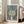 May include: Framed poster print leaning against white wall next to beige upholstered sofa in bright living room with large windows. Artwork features stylized tree with dotted circular canopy in greens and blues against swirling blue background with orange circles and white dots. Light wood frame, cream matting. French museum text at bottom. Natural lighting creates soft shadows on hardwood floor.