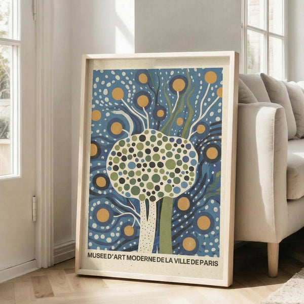 May include: Framed poster print leaning against white wall next to beige upholstered sofa in bright living room with large windows. Artwork features stylized tree with dotted circular canopy in greens and blues against swirling blue background with orange circles and white dots. Light wood frame, cream matting. French museum text at bottom. Natural lighting creates soft shadows on hardwood floor.