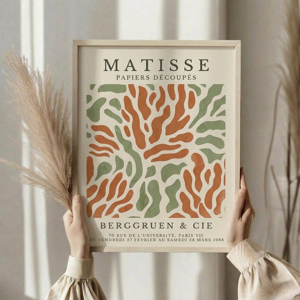 May include: Hands holding a framed art print featuring abstract organic shapes in sage green and terracotta orange on cream background, styled with dried pampas grass against soft neutral curtains in natural lighting.