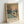 May include: Framed art print leaning against white wall showing winter waterside scene with bare trees, sailboats, and orange sun reflecting on water. Features impressionist painting style with blue, orange, and cream tones. Text below reads "ART CONTEMPORAIN INSPIRÉ PAR UN MAÎTRE ARTISTIQUE MONET" in black letters on cream background within light wood frame.