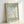 Framed poster print featuring a large white flower design on sage green background, displayed in light wood frame leaning against white wall. The artwork shows "MATISSE" at top and "PAPIERS DÉCOUPÉS" at bottom in gray text, with an abstract daisy-like bloom created from cut paper shapes in Matisse's signature style. The white petals have subtle texture and weathered appearance against the muted green backdrop.