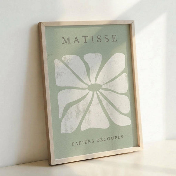 Framed poster print featuring a large white flower design on sage green background, displayed in light wood frame leaning against white wall. The artwork shows "MATISSE" at top and "PAPIERS DÉCOUPÉS" at bottom in gray text, with an abstract daisy-like bloom created from cut paper shapes in Matisse's signature style. The white petals have subtle texture and weathered appearance against the muted green backdrop.