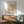 May include: Framed Matisse-inspired poster with orange and green organic cutout shapes on cream background, displayed above wooden sideboard with white ceramic lamp. Mid-century modern living room setting features sage green upholstered armchair with wooden frame, geometric wood coffee table, and warm natural lighting creating shadows on white wall.