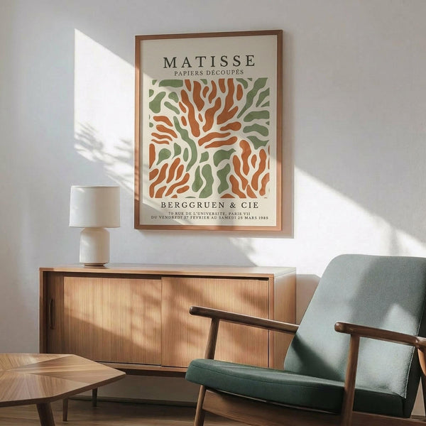 May include: Framed Matisse-inspired poster with orange and green organic cutout shapes on cream background, displayed above wooden sideboard with white ceramic lamp. Mid-century modern living room setting features sage green upholstered armchair with wooden frame, geometric wood coffee table, and warm natural lighting creating shadows on white wall.