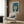 May include: Framed vintage-style art poster featuring stylized tree with dotted green canopy and flowing blue background with orange circles, displayed above beige upholstered armchair with geometric patterned pillow. Modern reading nook with brass floor lamp, dark round side table with open book, and warm wood paneled wall.